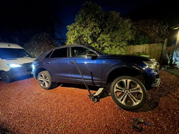Blue SUV raised on a jack during a nighttime mobile tyre fitting, with service tools beside the front wheel.