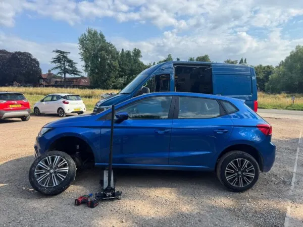 Blue SUV raised on a jack during mobile tyre fitting, with the front wheel removed and service tools beside the vehicle.