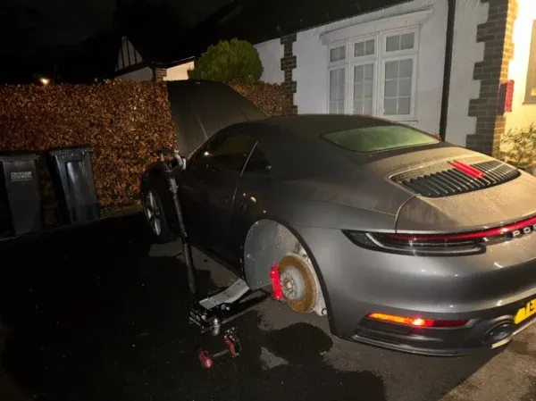 Grey Porsche raised on a jack during a nighttime mobile tyre change, with the rear wheel removed outside a residential property.