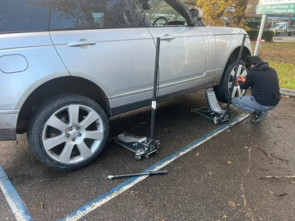 Mobile tyre technician changing a wheel on a silver Range Rover using professional jacks in a parking area.